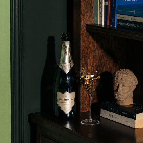 A bottle of Blanc De Blanc stands on a dark wooden cabinet beside a filled English Sparkling Wine flute against a dark green wall and wood shelving.