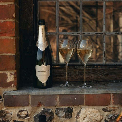 A bottle of Hattingley Blanc de Blanc sparkling wine and two filled English Sparkling Wine flutes resting on a stone windowsill beside a brick wall.