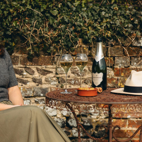 Bottle of Hattingley Valley Blanc de Blancs and two glasses on a table with a person sitting nearby, against a stone wall and greenery.