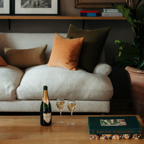 A styled living room with a light beige sofa, several throw pillows, a bottle of classic reserve, two filled champagne glasses on a wooden table, and a book in the foreground.