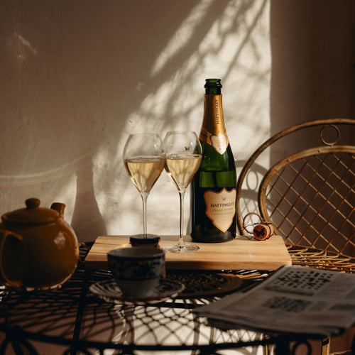 A bottle of Hattingley's Classic Reserve with two English Sparkling Wine flutes on a wooden board, styled on a table with a teapot, cup, newspaper, and woven chair in warm sunlight.