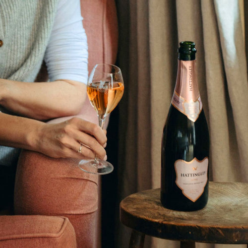 Bottle of Hattingley Valley Rosé English sparkling wine on a wooden stool beside a pink armchair. In the armchair a lady holds a glass of pink English Sparkling. With a blurred indoor background and linen curtain in the background.