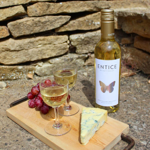 A bottle of Entice by Hattingley Valley, English dessert wine Bacchus. The bottle stands on a wooden board beside two filled wine glasses, a wedge of blue cheese, and a bunch of red grapes, set outdoors against a stone wall.
