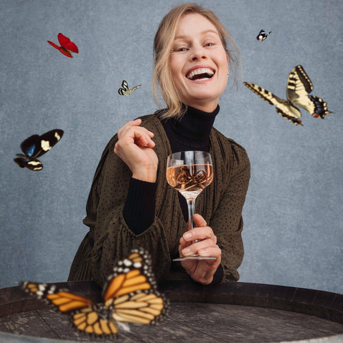 Lifestyle image of woman holding a glass of bubbles, laughing as she is surrounded by butterflies