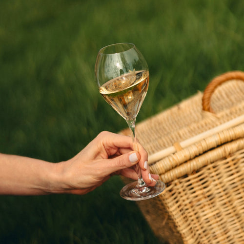 Image of a hand holding a Hattingley glass flute, the flute is filled with English sparkling wine and has the Hattingley logo with a shiny gold butterfly on it, in the background is spring green grass and a picnic basket