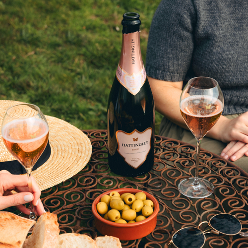 A bottle of Hattingley Rosé English Sparkling on an outdoor patio table, surrounded by two filled wine glasses, a bowl of olives, bread, sunglasses, and a straw hat. A person in a gray sweater is seated at the table.
