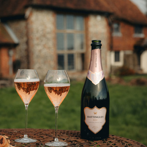 A bottle of Hattingley Rose with two English Sparkling Wine glasses set Infront of a rural country house.
