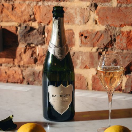 A bottle of Hattingley Blanc de Blancs with silver foil and label next to a full champagne flute on a marble counter with a brick wall background, lemons in the foreground