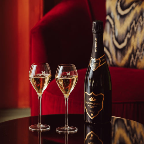 Bottle and two glasses of Hattingley Valley King's Cuvée English Sparkling wine on a reflective surface with a red velvet background.