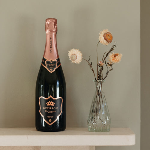 Bottle of Hattingley Valley Kings Rosé, prestige super premium English Sparkling Rosé on a white stone mantle with pink and white dried flowers in a glass vase beside the bottle, grey painted wall background.