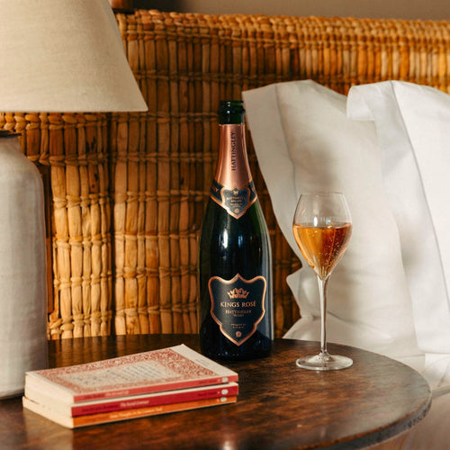 Bottle of Kings Rosé English Sparkling Wine and a glass on a wooden table with a lamp and books in the background.