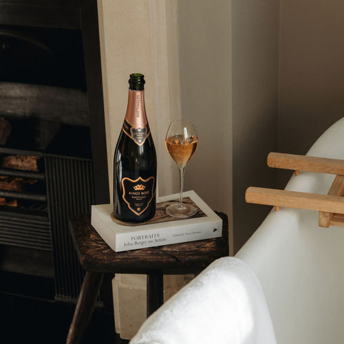 A bottle of Hattingley Valley Kings Rosé and filled glass on a white book placed on a stool beside a bathtub. English sparkling wine glass with pink wine fizzing, fireplace in the background.