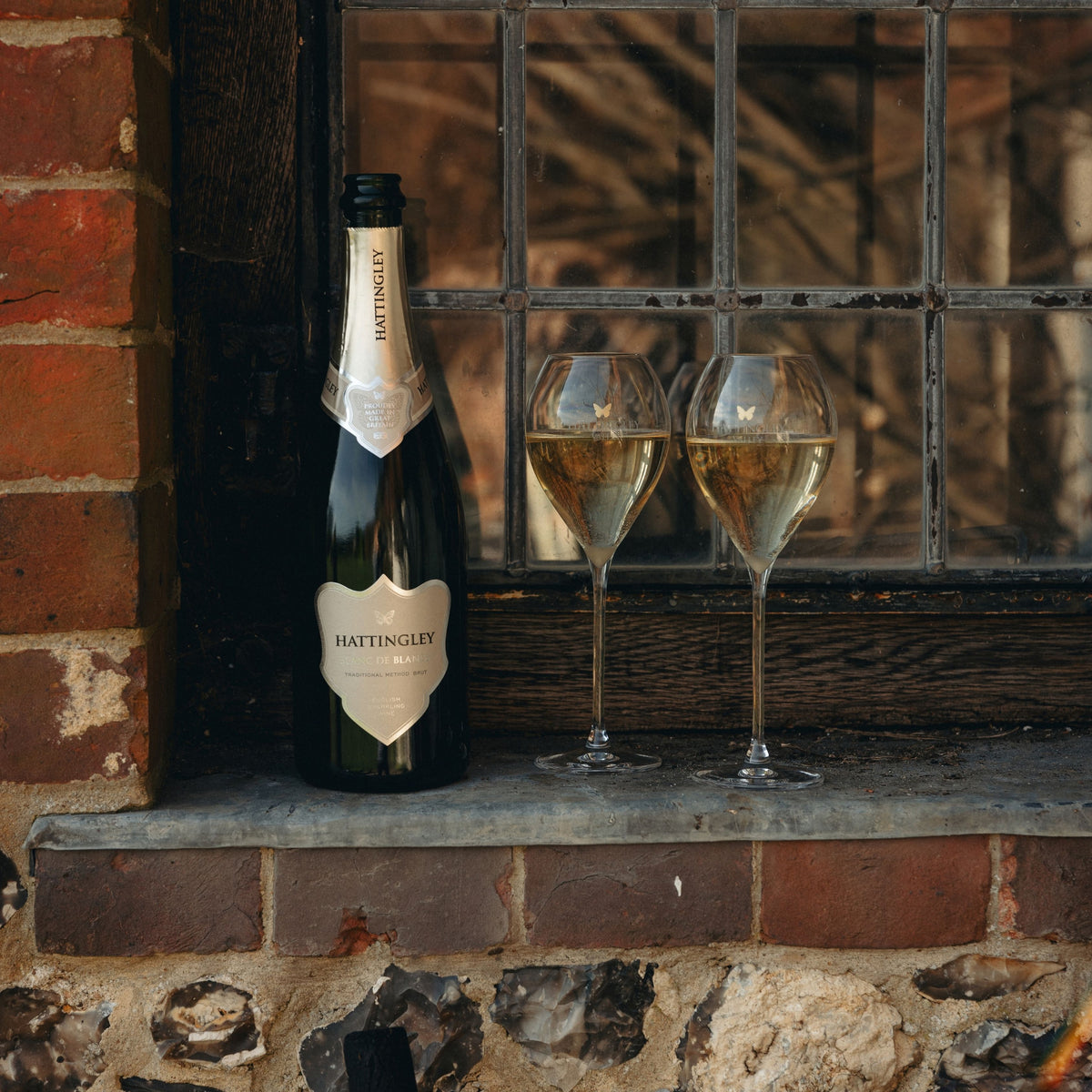 A bottle of Hattingley Blanc de Blanc sparkling wine and two filled English Sparkling Wine flutes resting on a stone windowsill beside a brick wall.