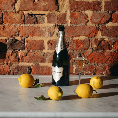 Bottle of Hattingley Valley Blanc de Blancs with a filled glass and lemons on a marble surface against a brick wall from far away. Silver label and foil on bottle.