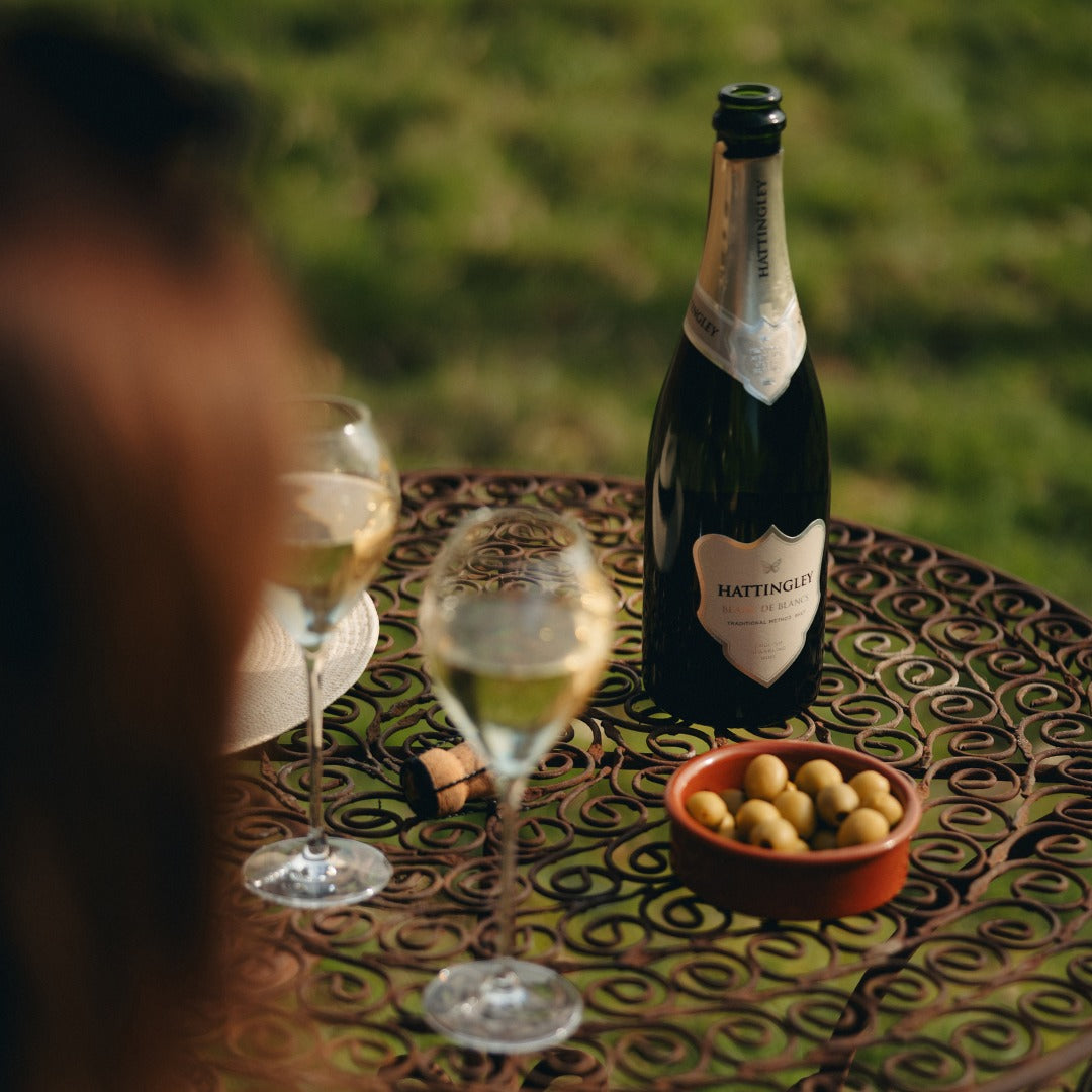 Bottle of Hattingley English Sparkling wine champagne with two glasses and a bowl of olives on a table outdoors.