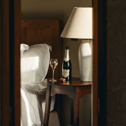 A bottle of Hattingley Valley award winning blanc de blancs wine with a silver label sat on a mahogany bedside table with a elegant glass flute filled with sparkling wine beside, a bed with white sheets in the background.