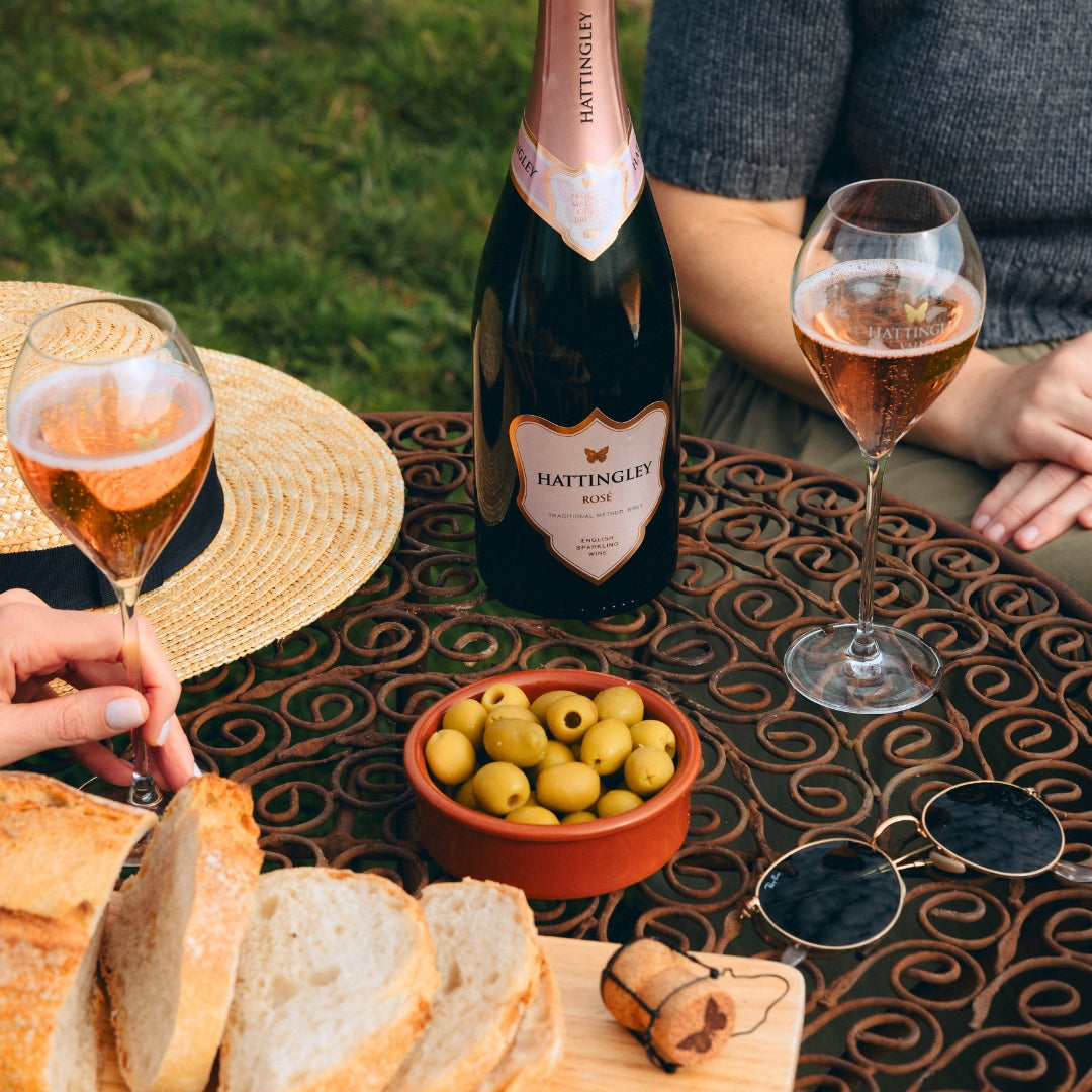 Two people enjoying a bottle of Hattingley Rosé with bread and olives outdoors. Close up shot on a metal table with sunglasses and a hat resting beside the glasses.