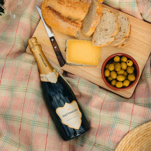 Bottle of Hattingley Valley sparkling wine with bread, cheese, and olives on a wooden board on a check picnic blanket