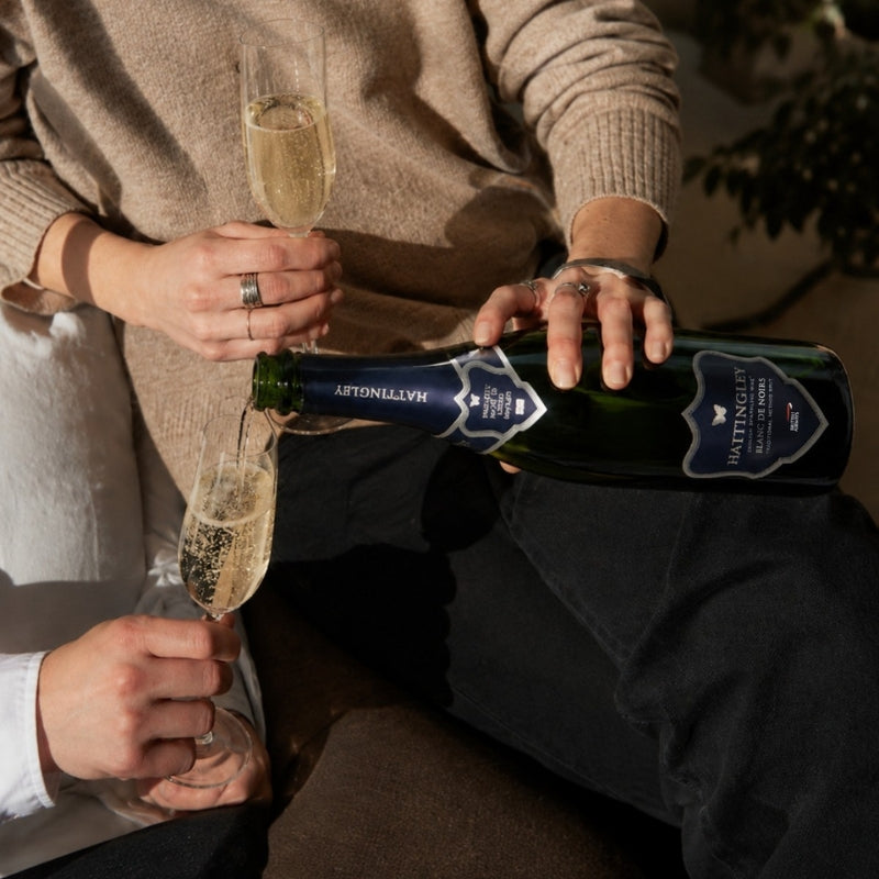 A lady in a beige jumper pours a bottle of British Airways Blanc de Noirs English Sparkling Wine from producer Hattingley Valley. Another hand holds out their glass flute to be poured. 