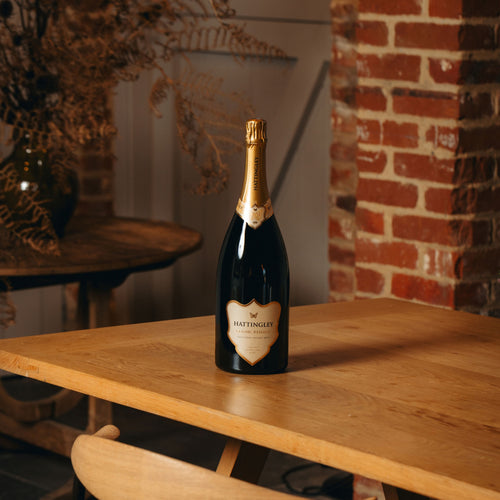 A bottle of Hattingley Classic Reserve Magnum stands on a light wood table in a warm, rustic interior setting with a brick wall and dried ferns in the background.