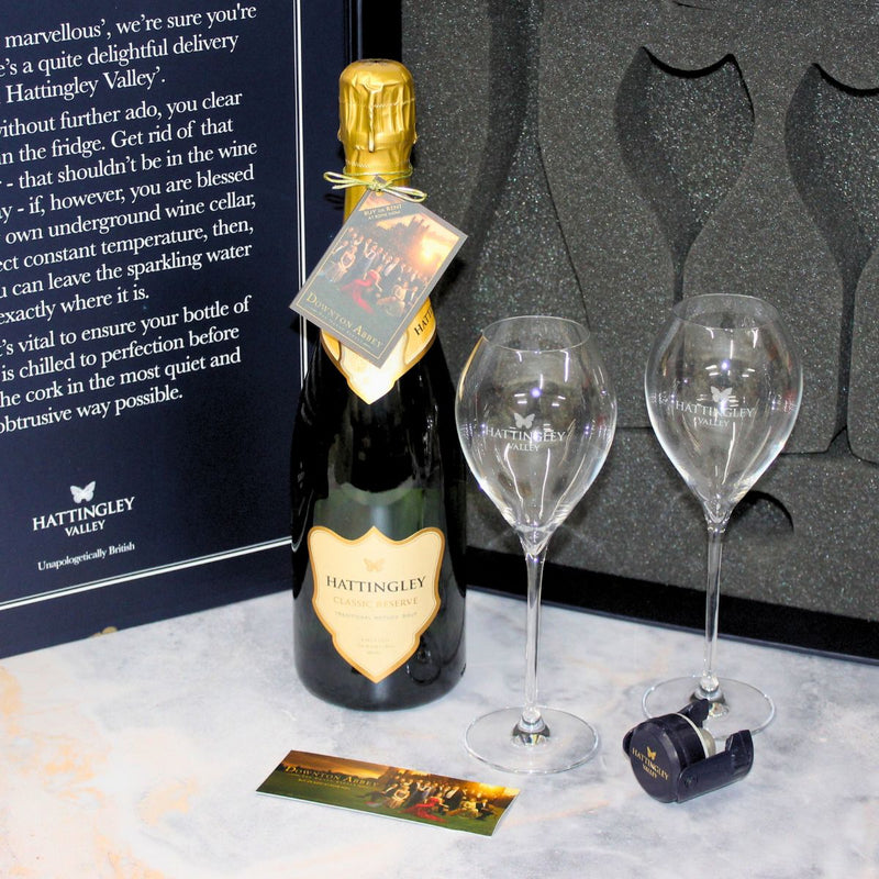 Open Luxury Gift set on a marble counter top with a pair of Hattingley valley branded tulip flutes and a sparkling wine bottle stopper in blue, included is a invitation to watch the new movie Downton Abbey: The Grand Finale at home. 