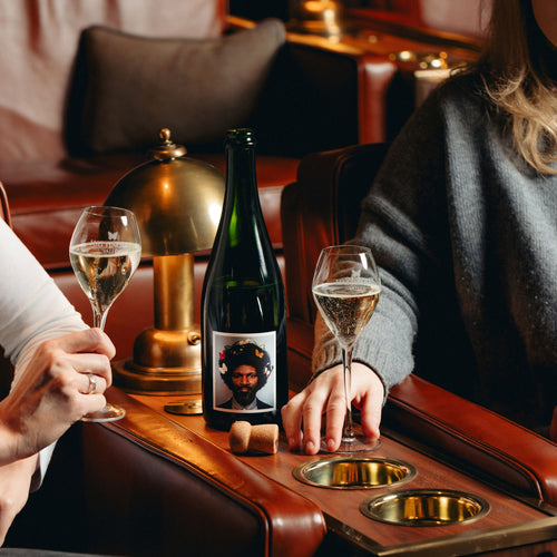 English Gent Bottle styled on a wooden table with two English Sparkling Wine flutes, a brass lamp, and a person in a grey sweater seated nearby.