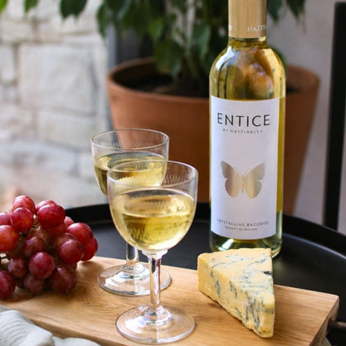 A bottle of Entice English dessert wine with two filled wine glasses, a wedge of blue cheese, and a bunch of red grapes arranged on a wooden board in a styled setting.