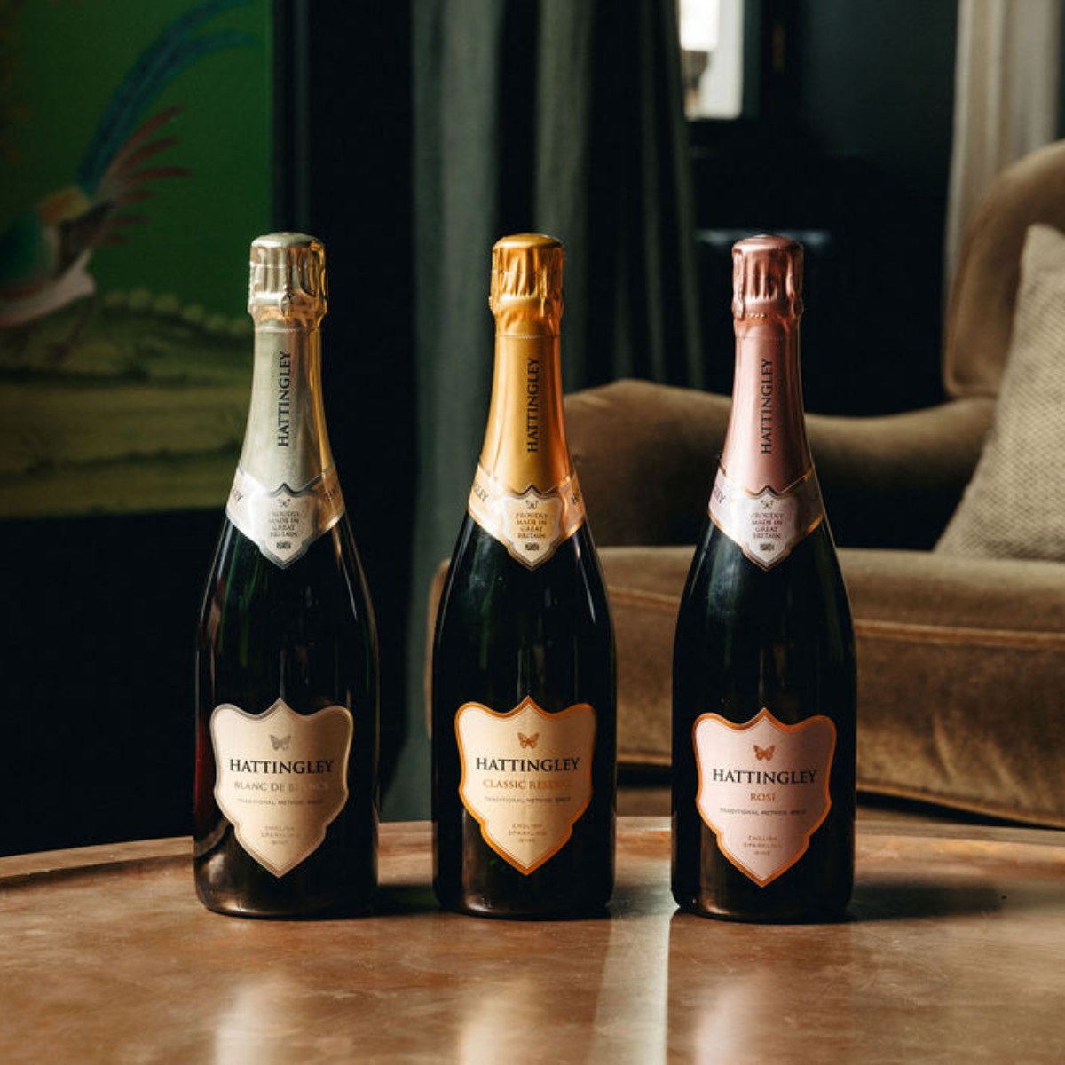 Three bottles of Hattingley Valley English sparkling wine on a wooden surface with a blurred background. The Core Collection including (left to right) Blanc de Blancs (with a silver label), Classic Reserve (with a gold label and Rosé Sparkling (with a pink label). Well lit with green wallpaper in the background.