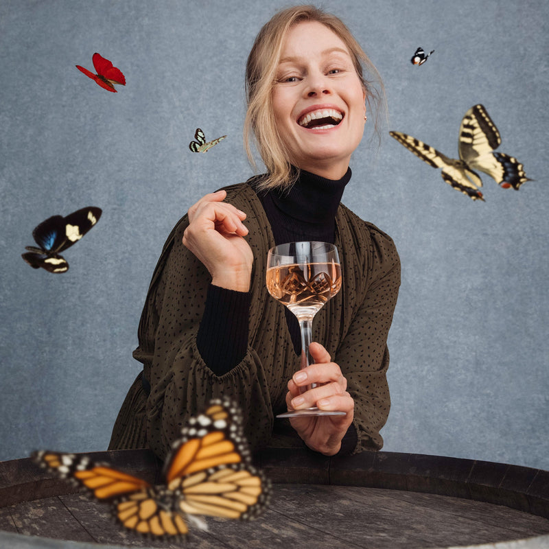 Lifestyle image of woman holding a glass of bubbles, laughing as she is surrounded by butterflies 