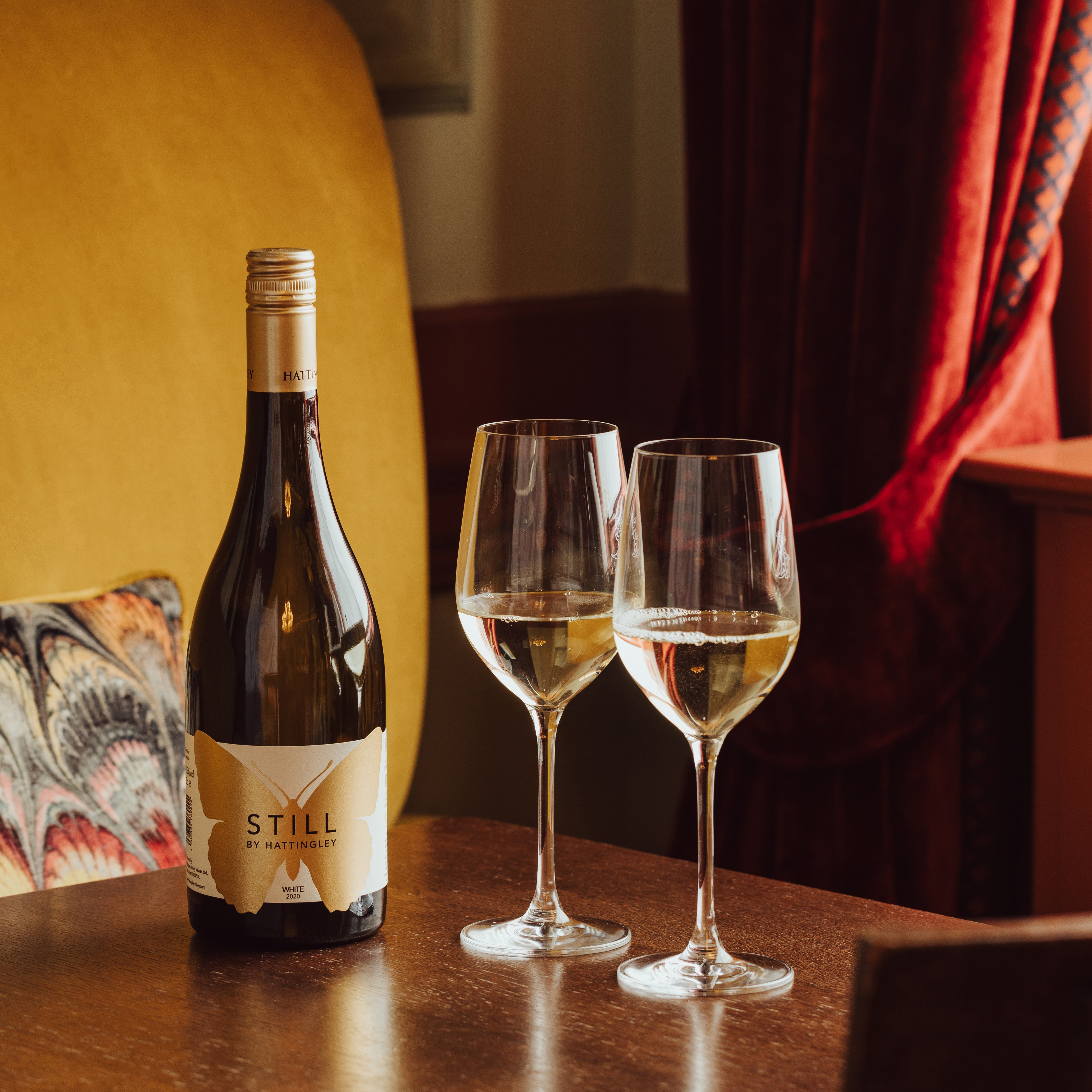 Hattingley Valley Still White, lifestyle image two filled glasses and a bottle of wine on table, luxurious hotel background 
