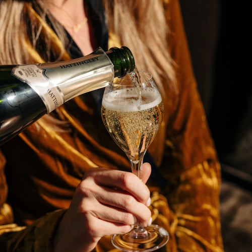 A close up lifestyle shot of a person pouring english sparkling wine into a flute glass, with the person wearing a shiny gold velvet robe.