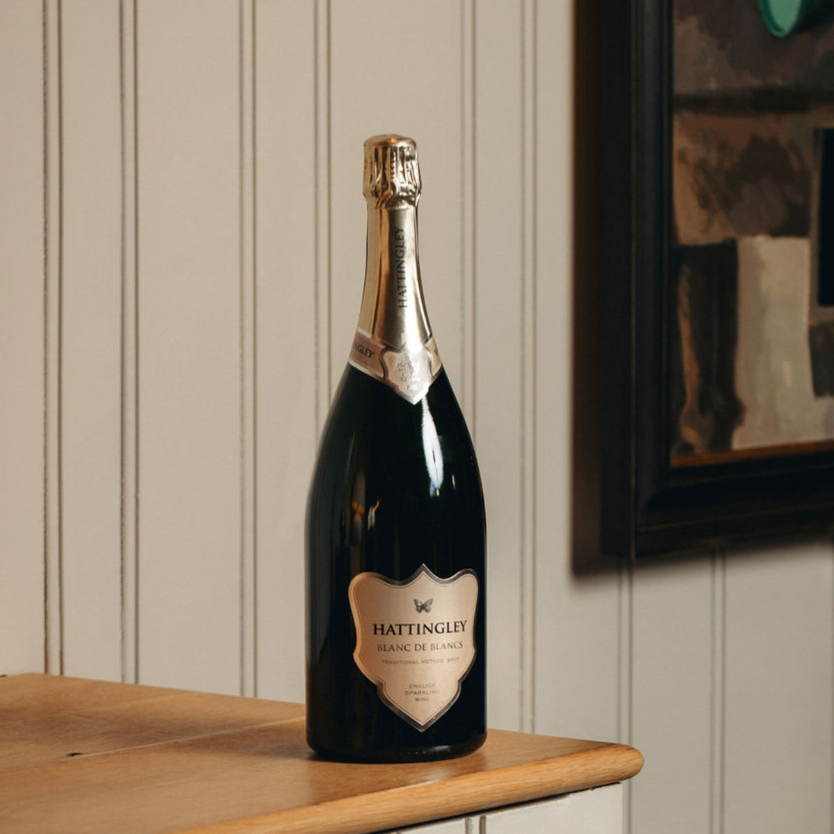 Hattingley Valley Magnum of Blanc de Blancs, large bottle of English Sparkling Wine With a silver label and foil on a wooden ledge with a slatted painted background in grey / green.