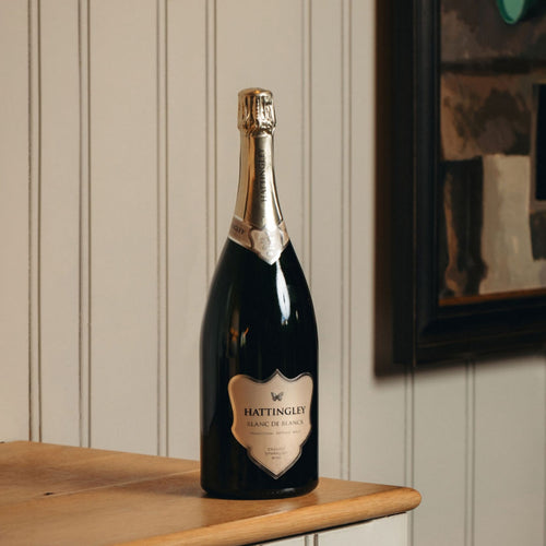 Hattingley Valley Magnum of Blanc de Blancs, large bottle of English Sparkling Wine With a silver label and foil on a wooden ledge with a slatted painted background in grey / green.