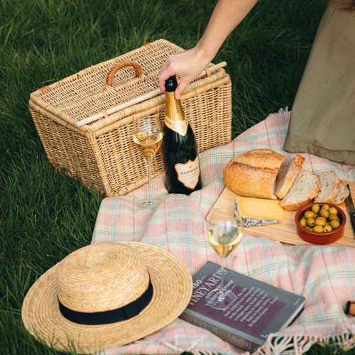 Picnic setup with a wicker basket, Hattingley Valley English Sparkling Wine, bread, and cheese on a checkered blanket outdoors.