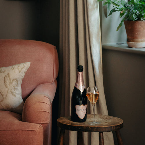 A bottle of Hattingley Rose and a filled English sparkling wine flute on a small wooden side table next to a dusty rose armchair and beige curtains.