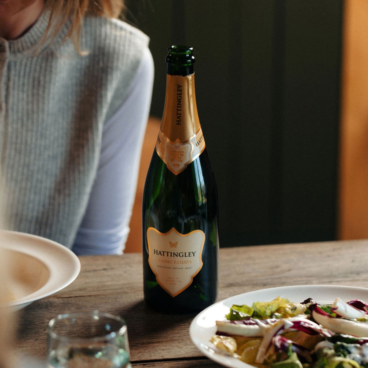 Bottle of Hattingley Classic Reserve English sparkling wine on a wooden table with plates and salad. The bottle has a gold foil and label and is the centre of the frame a lady sits in the background.