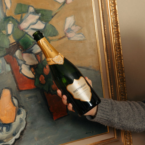 Hand holding a bottle of Hattingley Valley Classic Reserve English Sparkling wine (champagne method) in front of a gilded framed painting of abstract flowers with a gold background. The bottle itself has a gold label and gold foil.