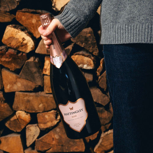 Person holding a Magnum bottle of Hattingley Valley Rose English Sparkling Wine against a background of stacked firewood. The Magnum of Rosé has a pink label and top foil.