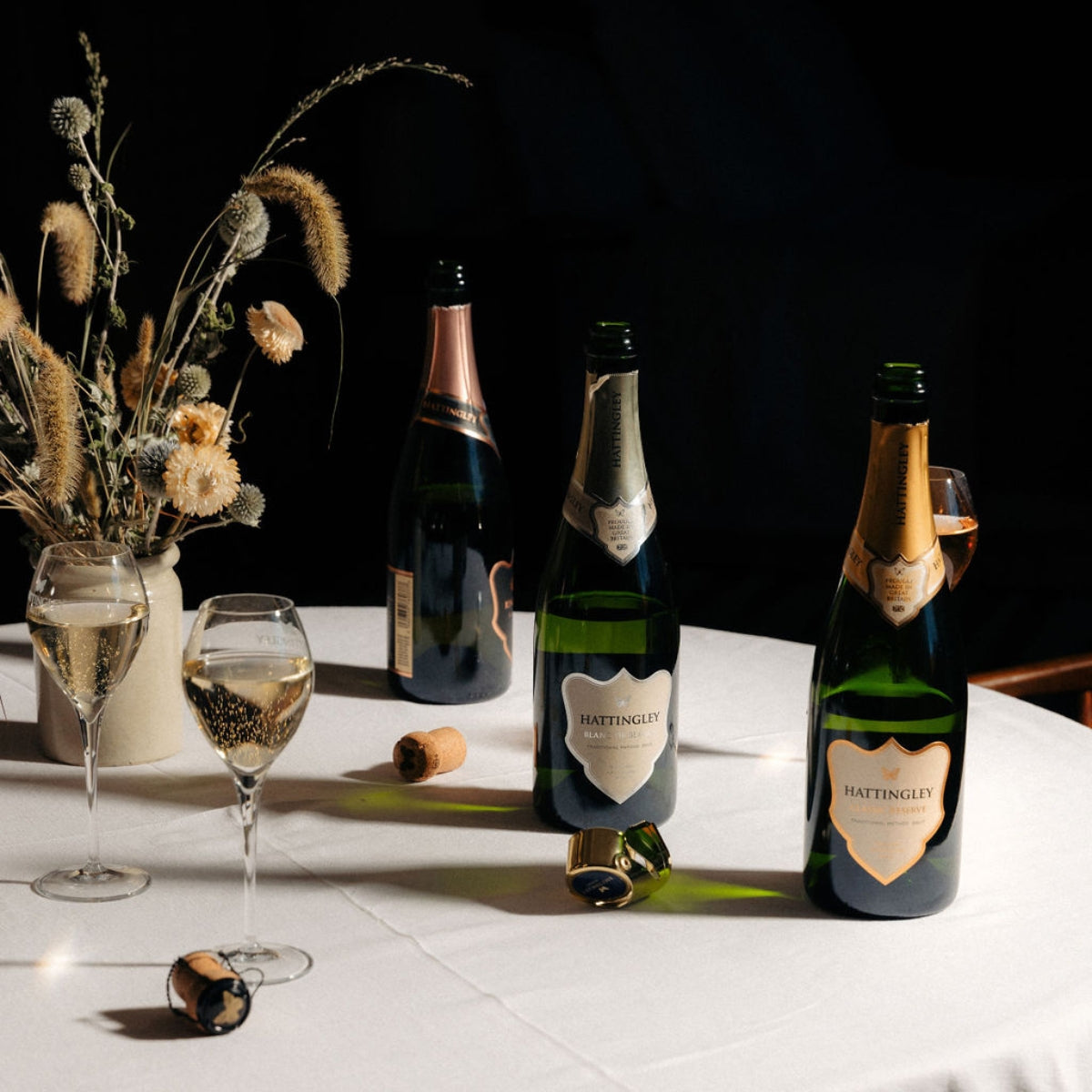 Hattingley English sparkling wine bottles on a table with a white table cloth glasses and flowers and a dark background.
