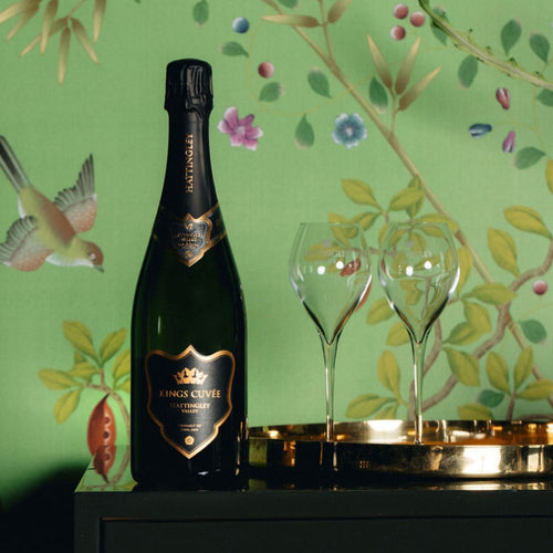Bottle of Kings Cuveé English Sparkling wine with two glasses on a decorative table against a floral wallpaper background. Black and gold label super premium English Wine, prestige cuvée from Hattingley Valley.