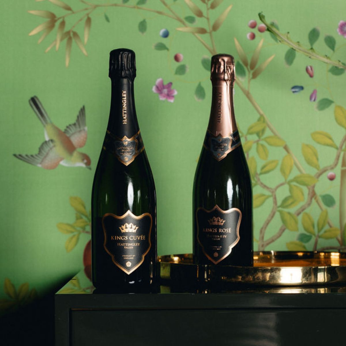 Two bottles of English Sparkling Wines on a gold tray with a decorative green wallpaper background. Kings Cuvée and Kings Rosé super premium English Sparkling wines by Hattingley Valley