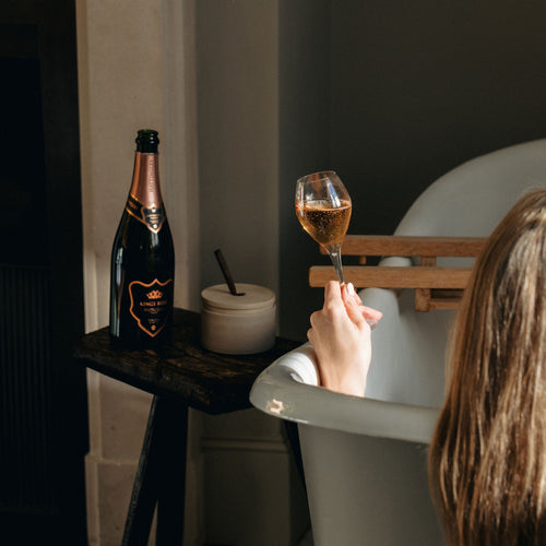 A person sitting in a bathtub holding a glass of English Sparkling Wine. To the left of the tub, a bottle Hattingley Kings Rose and candle sits on a small wooden table.