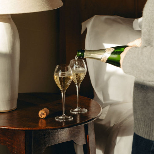 A lady pours two glass flutes of Hattingley Valley Blanc de Blanc sat on a bedroom side table with white pillows in the background.