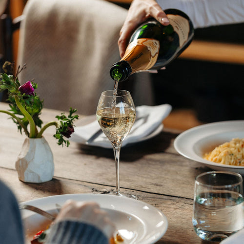 Person pouring Hattingley Classic Reserve English Sparkling wine into a glass on a dining table with plates and a vase of flowers.