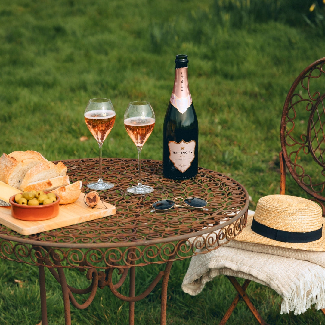 Bottle of English Sparkling Wine Rosé, two glasses, and a picnic setup on a metal table outdoors.