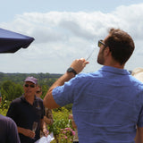 Man in blue shirt sipping a glass of English Sparkling Wine in the vineyard with a clear sky and trees in the background