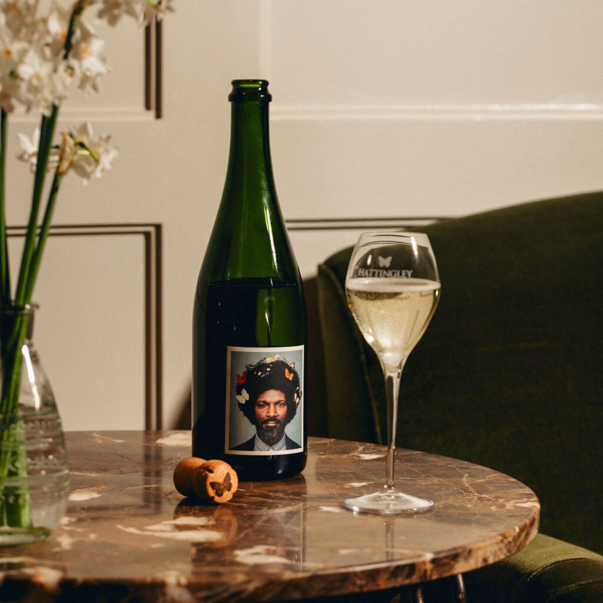 The English Gent, Bacchus and Pinot Gris blend traditional method English Sparkling Wine on a marble table with a filled champagne glass beside and a bottle cork with a butterfly symbol on the table. Also on the table is a small glass vase with white spring flowers in it.