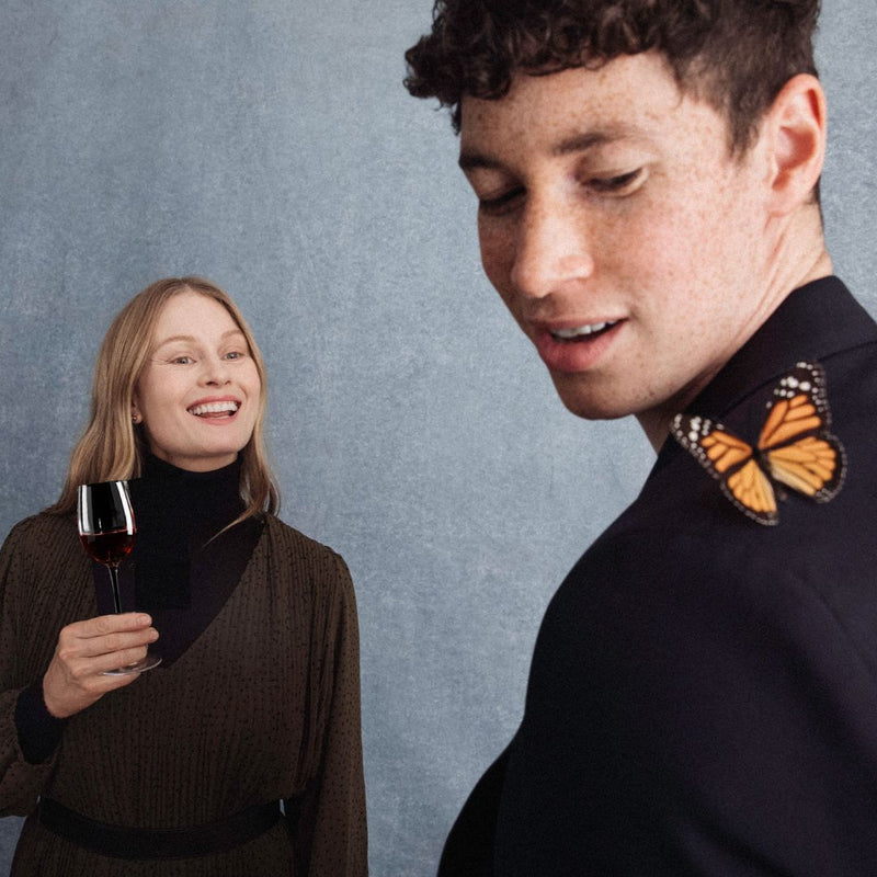 Hattingley Valley lifestyle image, a Lady holding a glass of red wine looks over laughing at a Gentleman with a butterfly on his shoulder. English Pinot Noir, English Red Wine. Best English Still Wine producers.