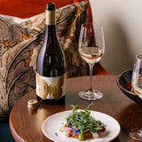A lifestyle image of Hattingley Valley STILL Chardonnay next to a glass of wine, with a plate of food in the foreground, against a luxury cushion in the background