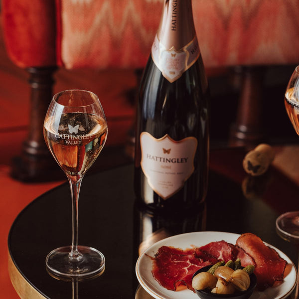 Hattingley Valley, Lifestyle Image by Rebecca Hope. Bottle of Hattingley Valley English Rosé on a reflective table with a filled glass and a food pairing of charcuterie, in the background is are luxurious hotel furnishings